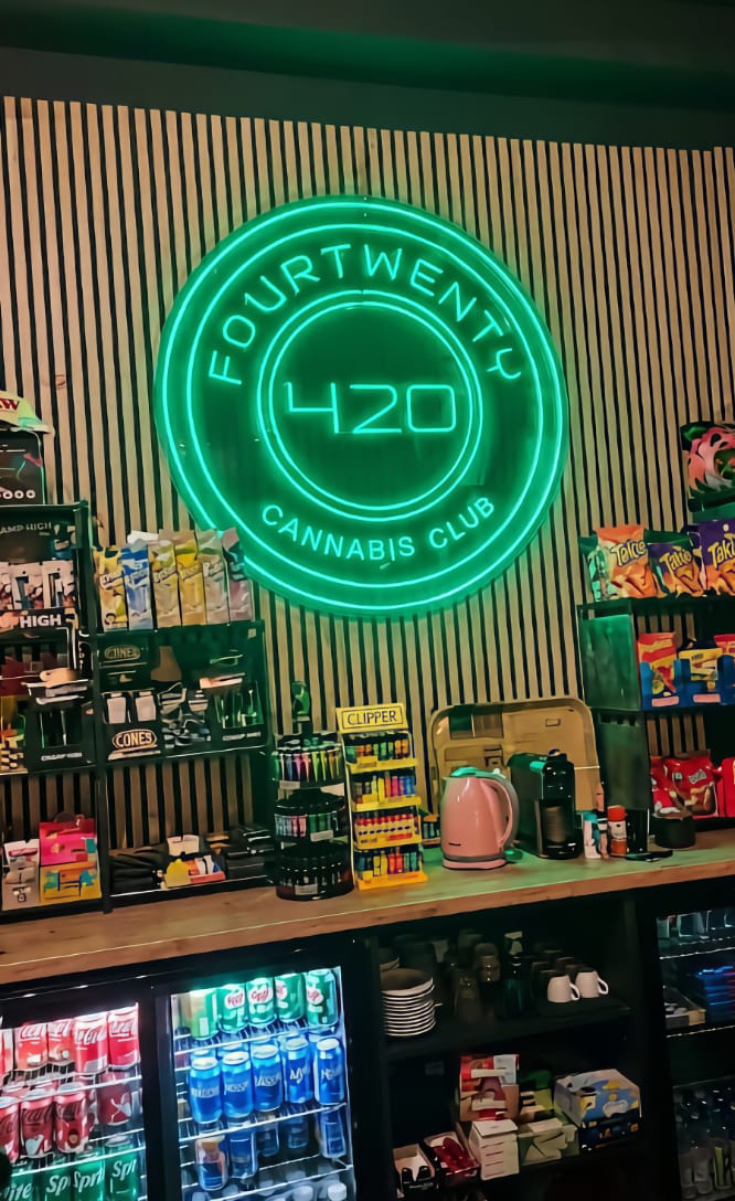 Ein hellgr&uuml;nes Neon-Schild mit dem Logo im Four Twenty Cannabis Club in Valencia, angebracht &uuml;ber einer Bartheke mit Snacks, Getr&auml;nken und Clipper-Feuerzeugen.