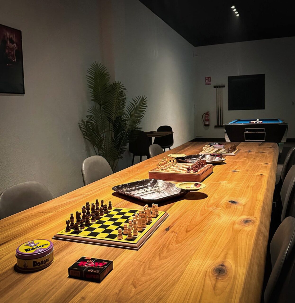 Ein langer Holztisch mit Schachbrettern, Brettspielen und Raucherzubeh&ouml;r im Four Twenty Cannabis Club in Valencia, mit einem Billardtisch im Hintergrund.
