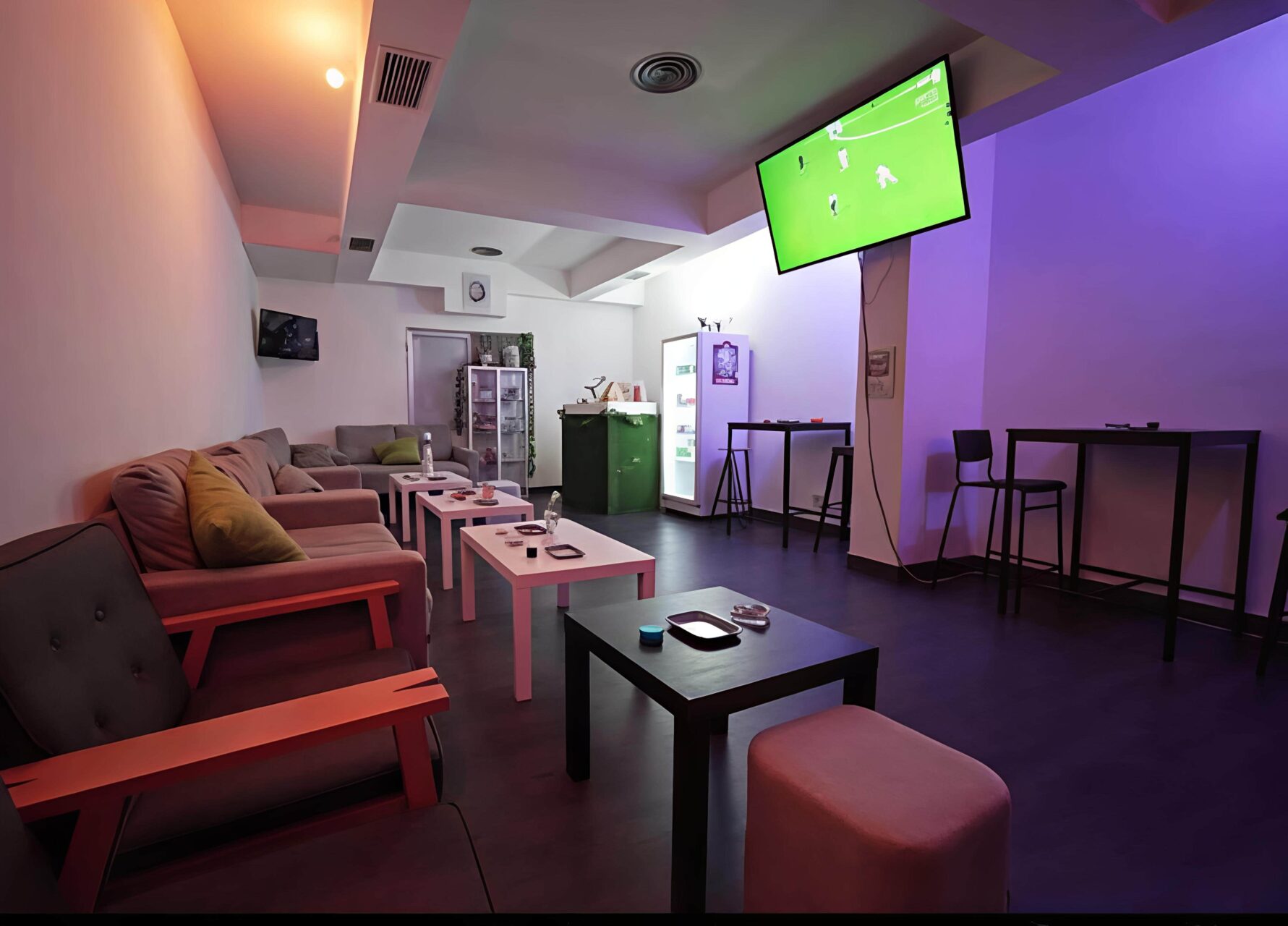 Lounge-Bereich im Valhalla Cannabis Club in Madrid mit Sitzgelegenheiten, Stehtischen, einem K&uuml;hlschrank und einem Fernseher, der ein Fu&szlig;ballspiel zeigt.