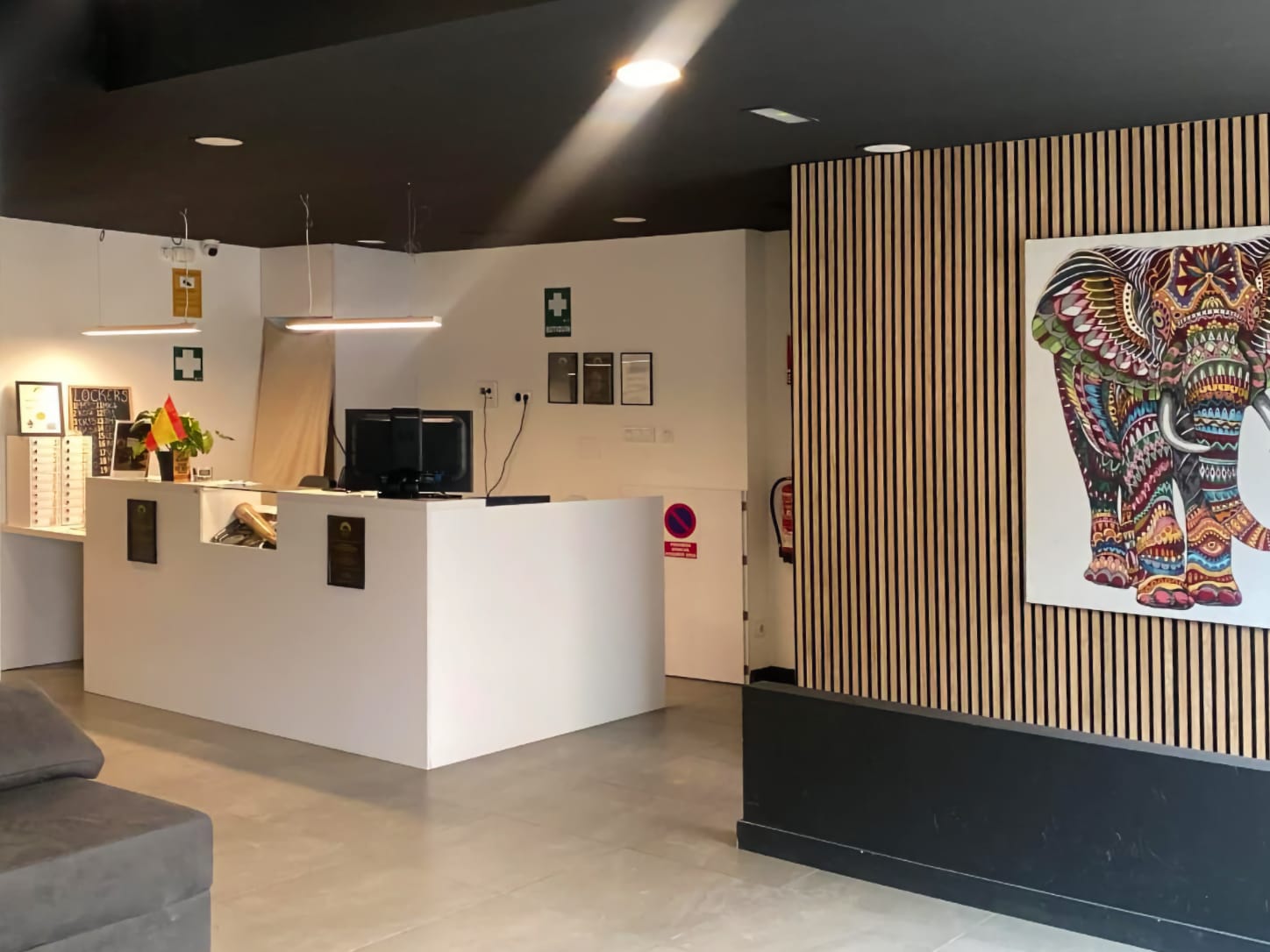 Die minimalistische wei&szlig;e Empfangstheke im Four Twenty Cannabis Club in Valencia, mit moderner Beleuchtung und einem farbenfrohen Elefantengem&auml;lde an einer Holzlamellenwand.